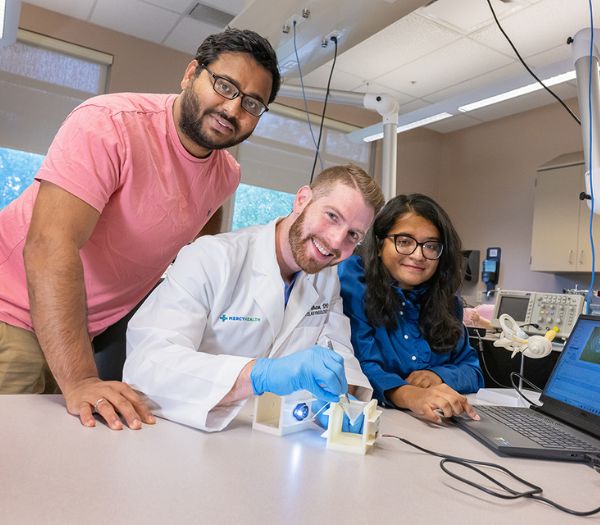 Three University of Akron students work together in a laboratory, smiling beside research equipment and a computer displaying experiment data.